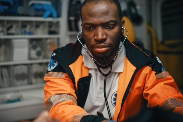 Close-up Portrait Shot of a Serious and Focused Black African American Paramedic in an Ambulance Vehicle with an Injured Patient. Emergency Medical Technician Uses Stethoscope to Monitor the Condition
